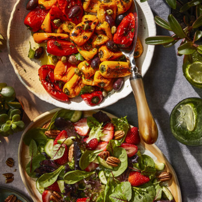 Two bowls of colorful salads: one with roasted red and yellow peppers, olives, and capers; the other with mixed greens, strawberries, pecans, and cucumber slices, surrounded by green plants and lime halves.